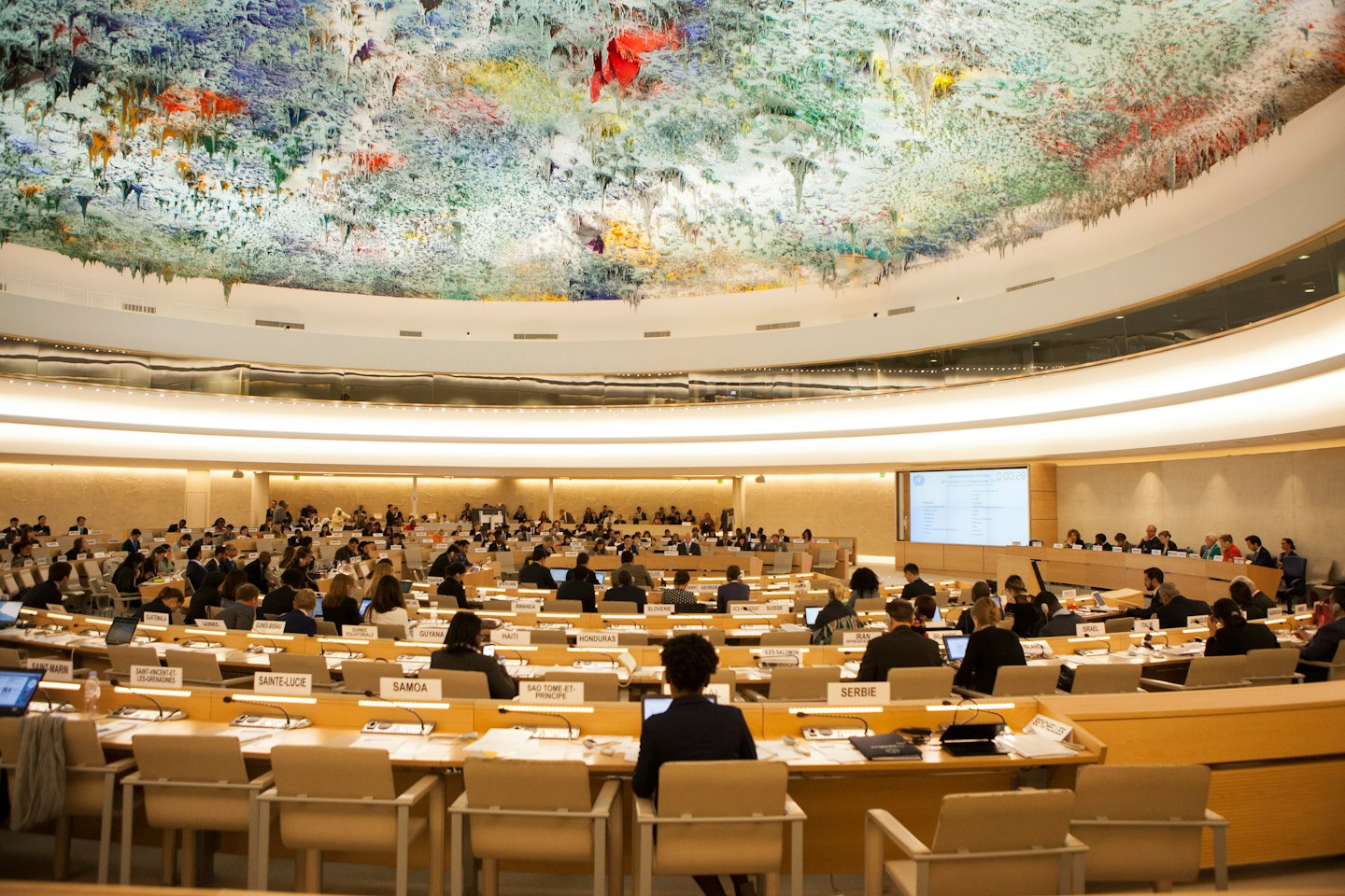 Delegates in a modern conference hall attend a meeting under a colorful domed ceiling.